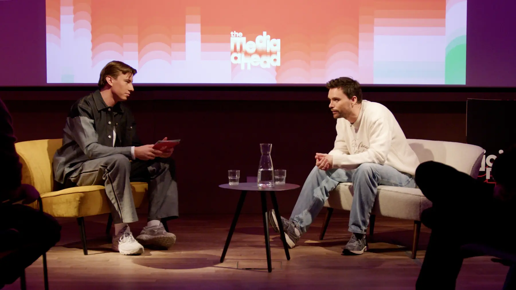 Twee mannen in gesprek op een podium met een groot scherm op de achtergrond.
