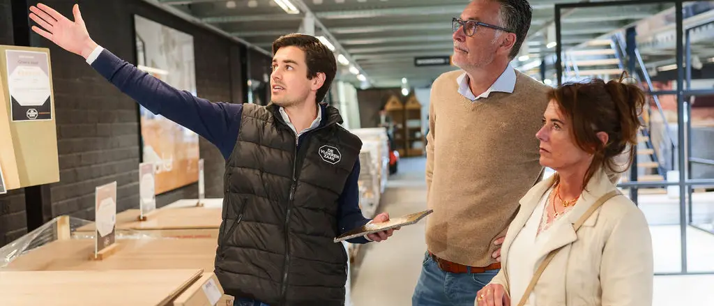 Een medewerker van De Vloerenzaak wijst een stel iets aan in de lichte showroom.