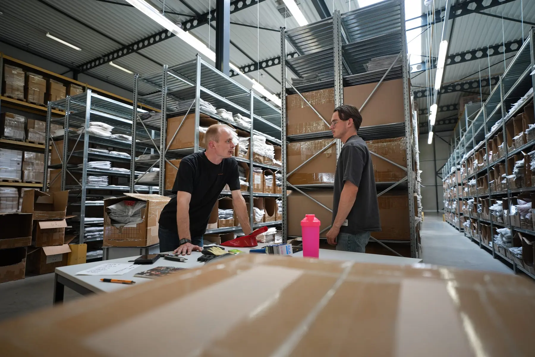 Twee medewerkers praten bij een werktafel in het magazijn.