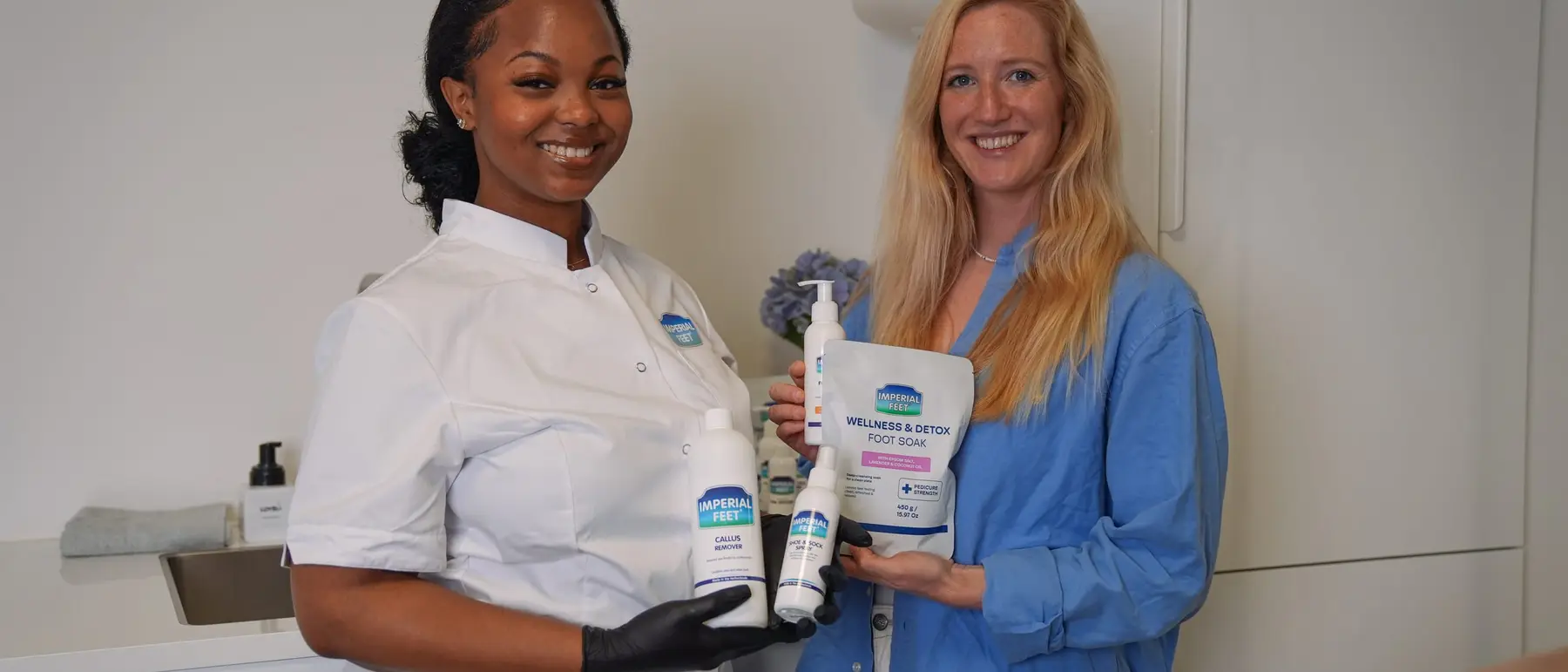 Een pedicure in witte jas en een vrouw in een blauw overhemd houden samen producten van Imperial Feet vast in een lichte salon.