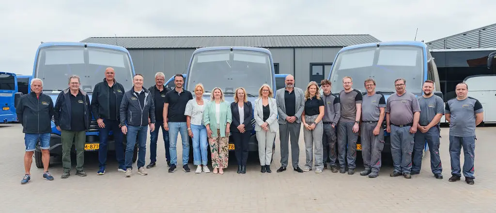 Een team staat in een lange rij voor drie bussen op een bedrijfsterrein.