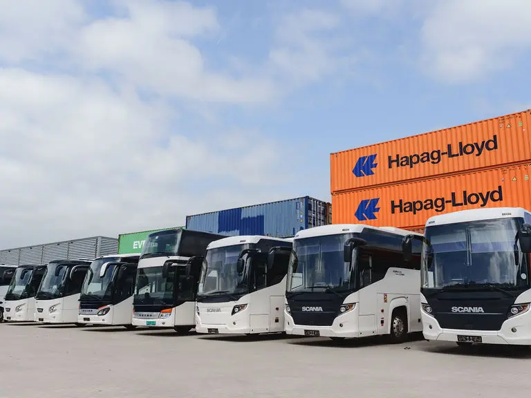 Een rij witte touringcars op een terrein, met gestapelde oranje containers erachter.