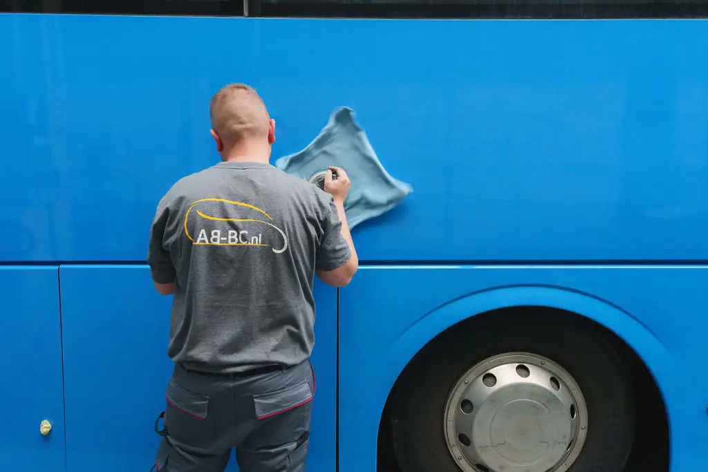 Een medewerker in AB-BC-shirt poetst de zijkant van een blauwe bus.