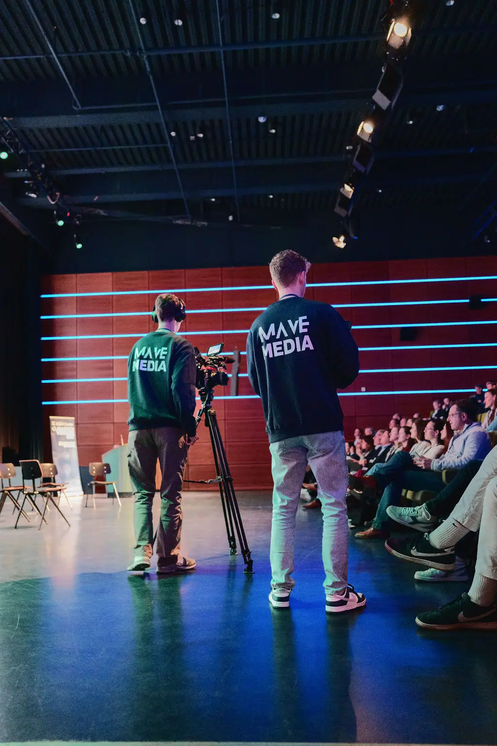 Twee cameramensen filmen een zaal vol publiek tijdens een presentatie.