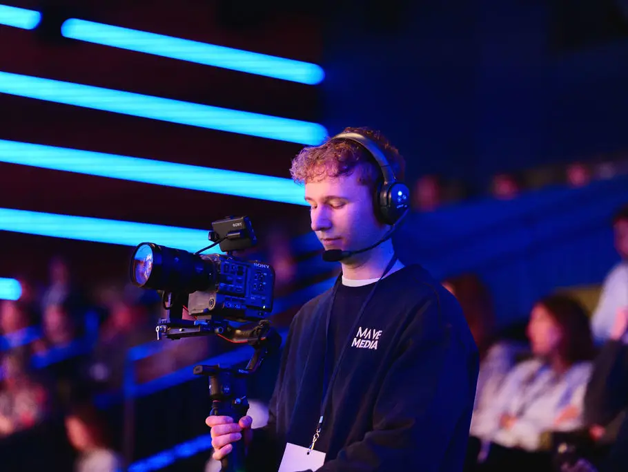 Een cameraman met koptelefoon filmt in een zaal met blauw licht en publiek.