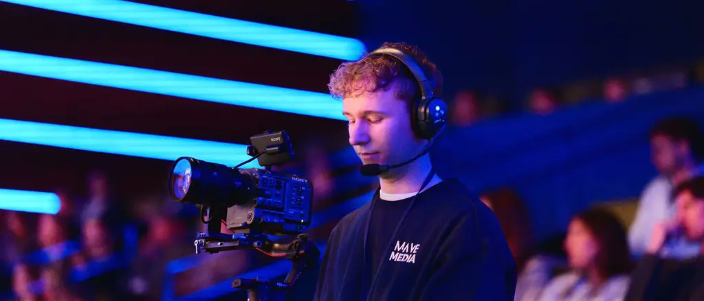 Een cameraman met koptelefoon filmt in een zaal met blauw licht en publiek.