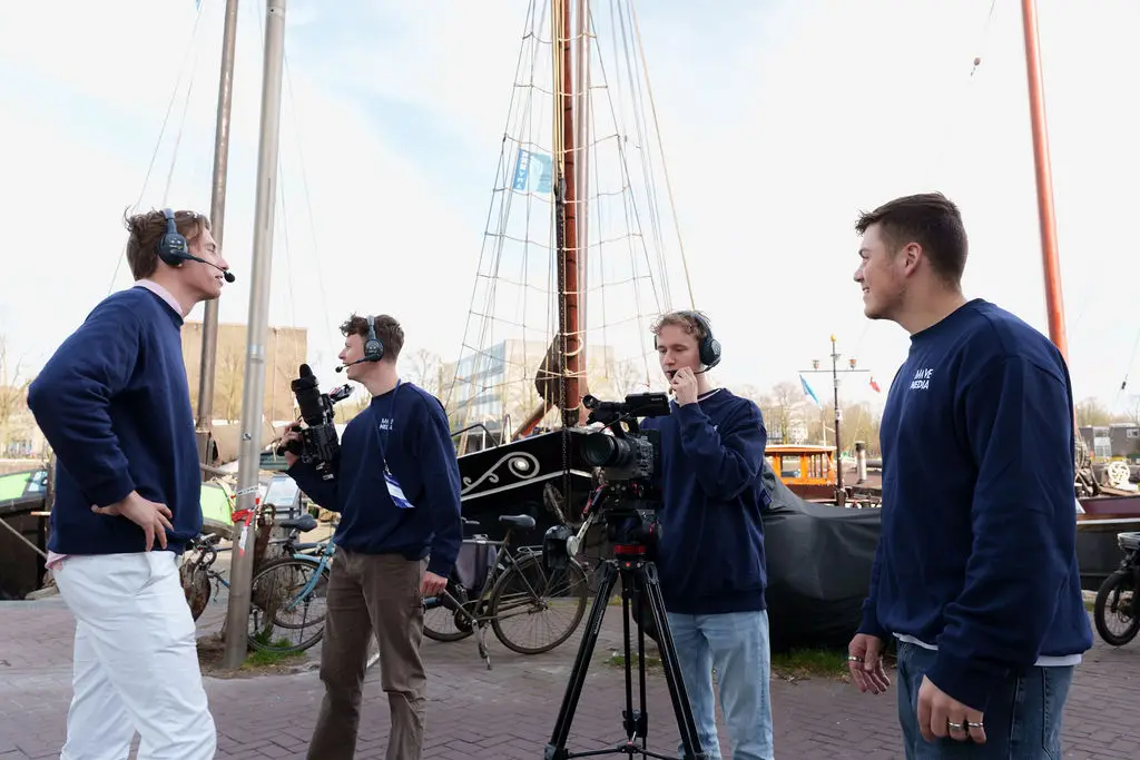 Een crew met camera op statief en koptelefoons aan een kade met oude schepen.