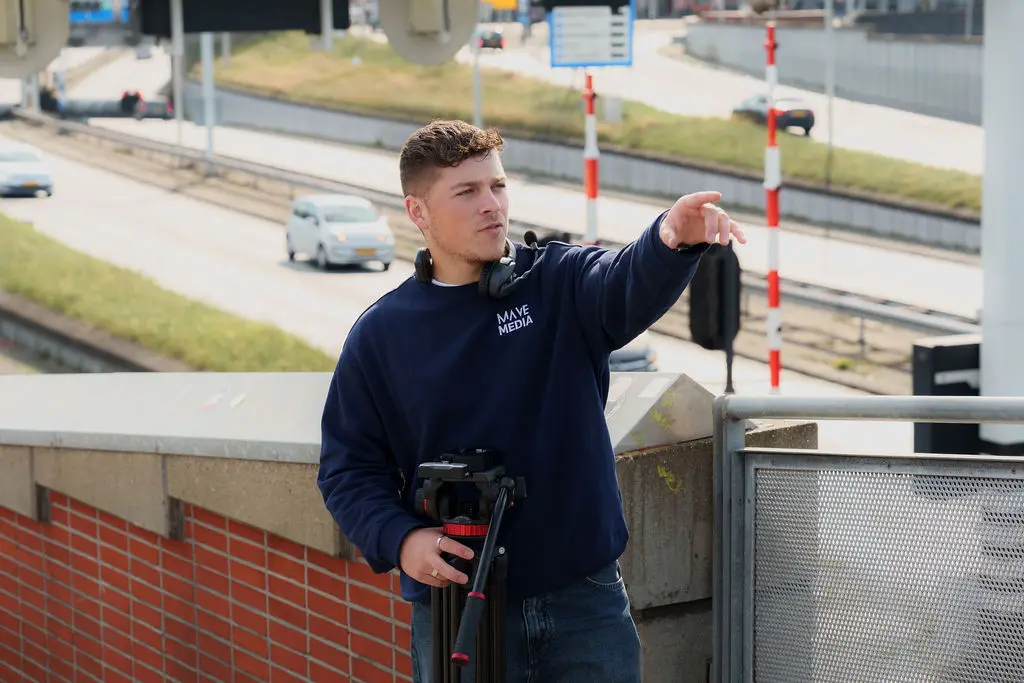 Een cameraman wijst iets aan, met een weg en slagbomen op de achtergrond.