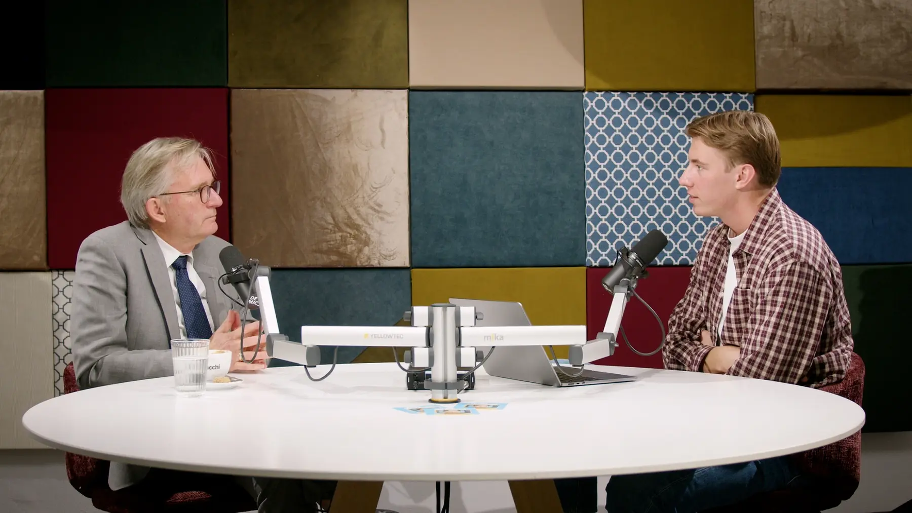 Een oudere man in pak en een jonge man in geruit overhemd in gesprek achter studiomicrofoons.