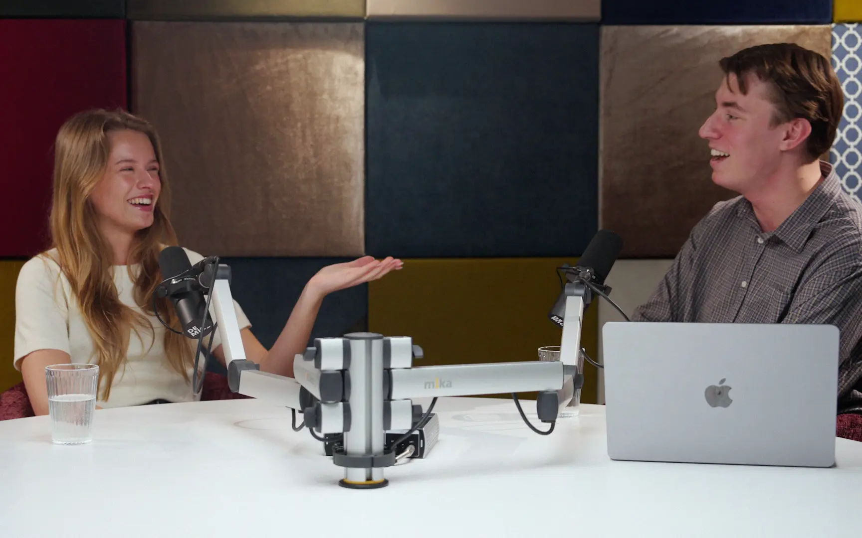 Een vrouw vertelt lachend iets aan een ronde studiotafel met microfoons.