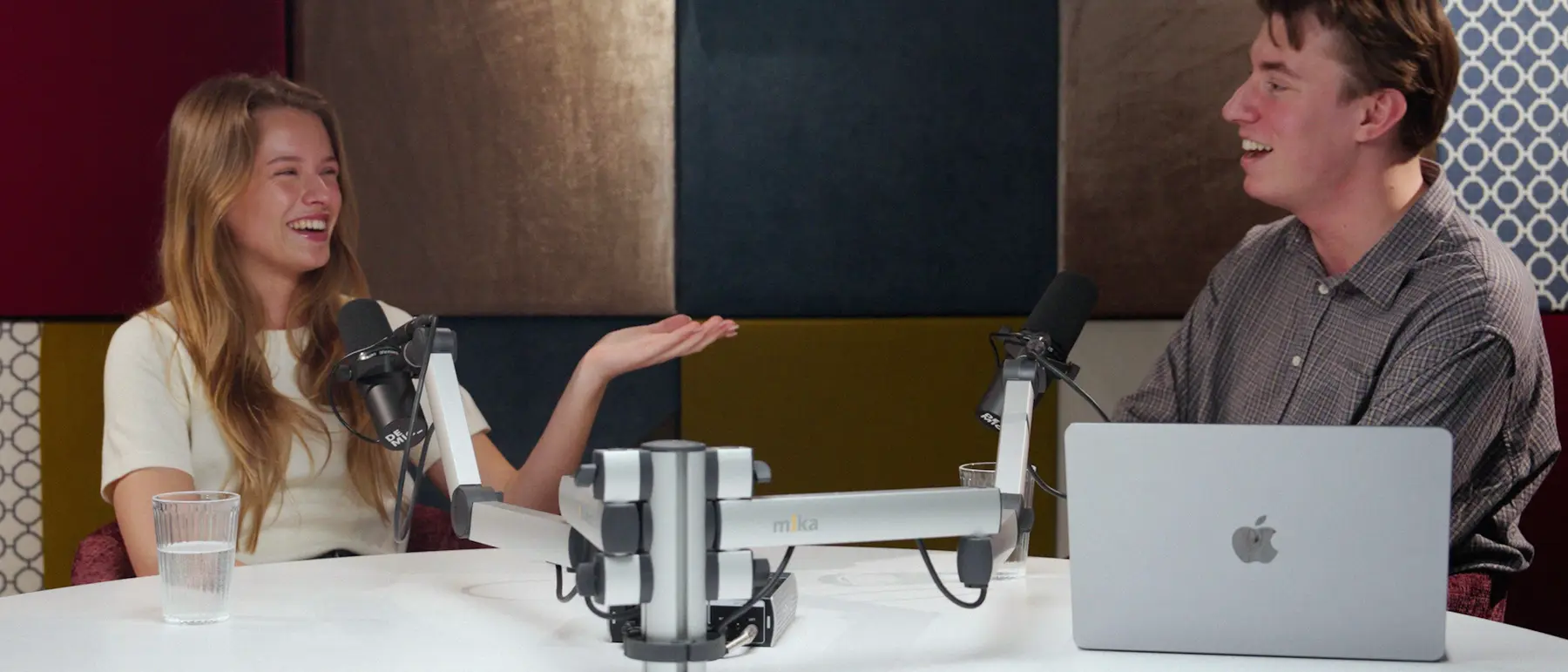 Een lachende vrouw vertelt iets aan een ronde studiotafel met microfoons, tegenover een jonge man.