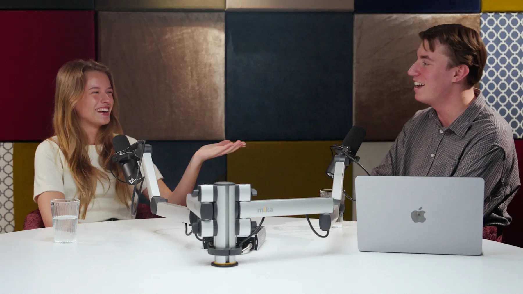 Een lachende vrouw vertelt iets aan tafel in de podcaststudio, tegenover een jonge man.