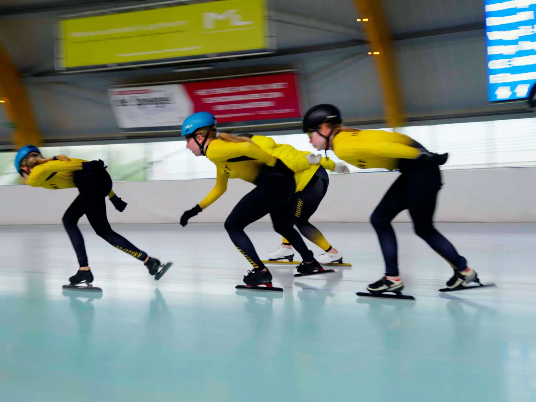 Drie shorttrackers in gele pakken nemen een bocht op de ijsbaan.