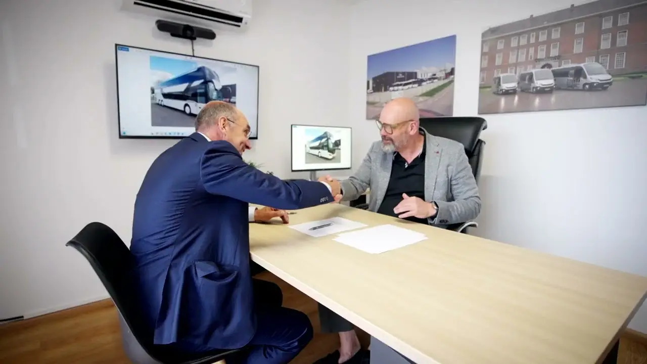 Twee mannen schudden elkaar de hand aan een bureau, met foto's van bussen op de achtergrond.