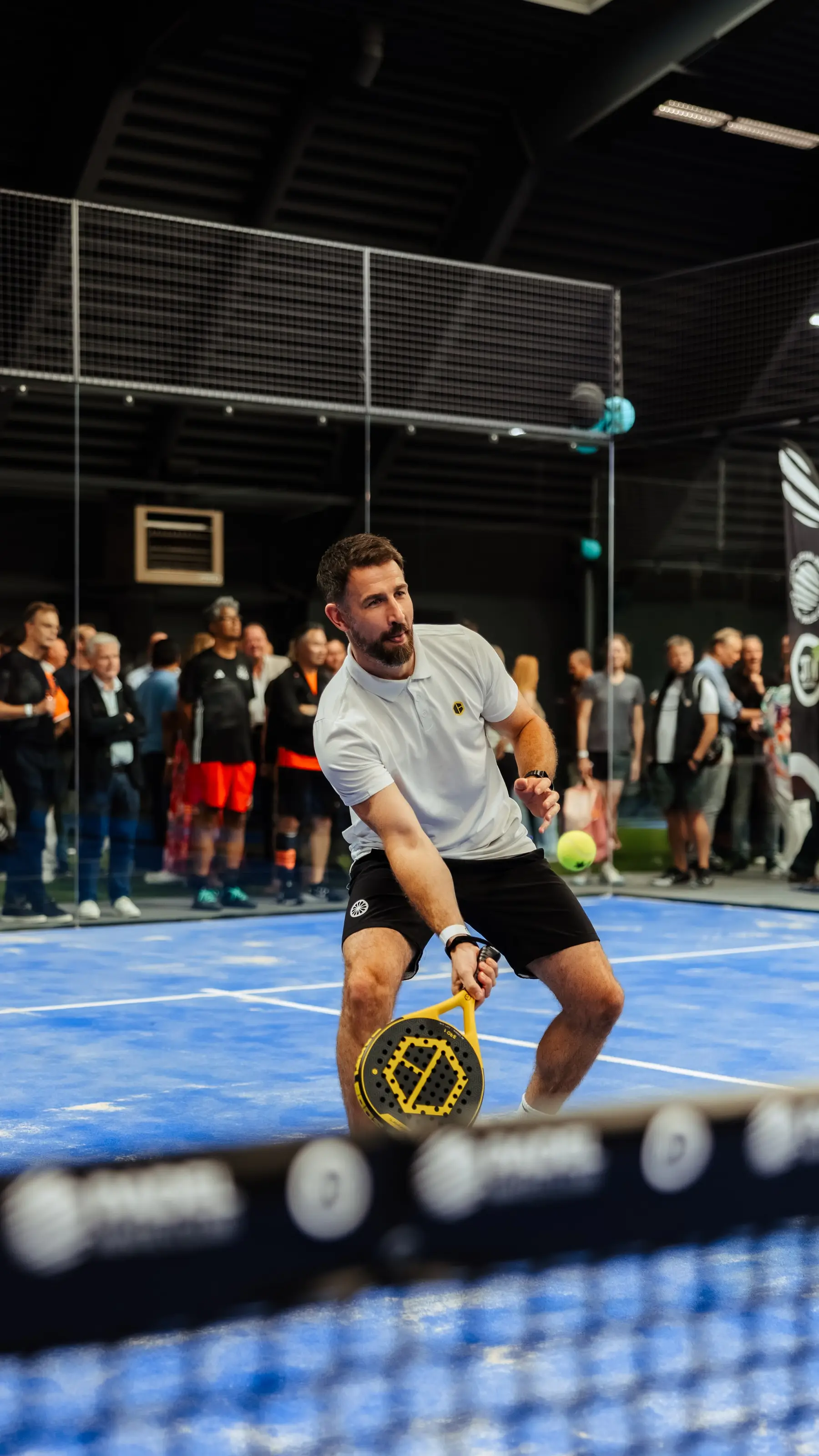 Een padelspeler speelt een bal voor publiek dat langs de baan staat.
