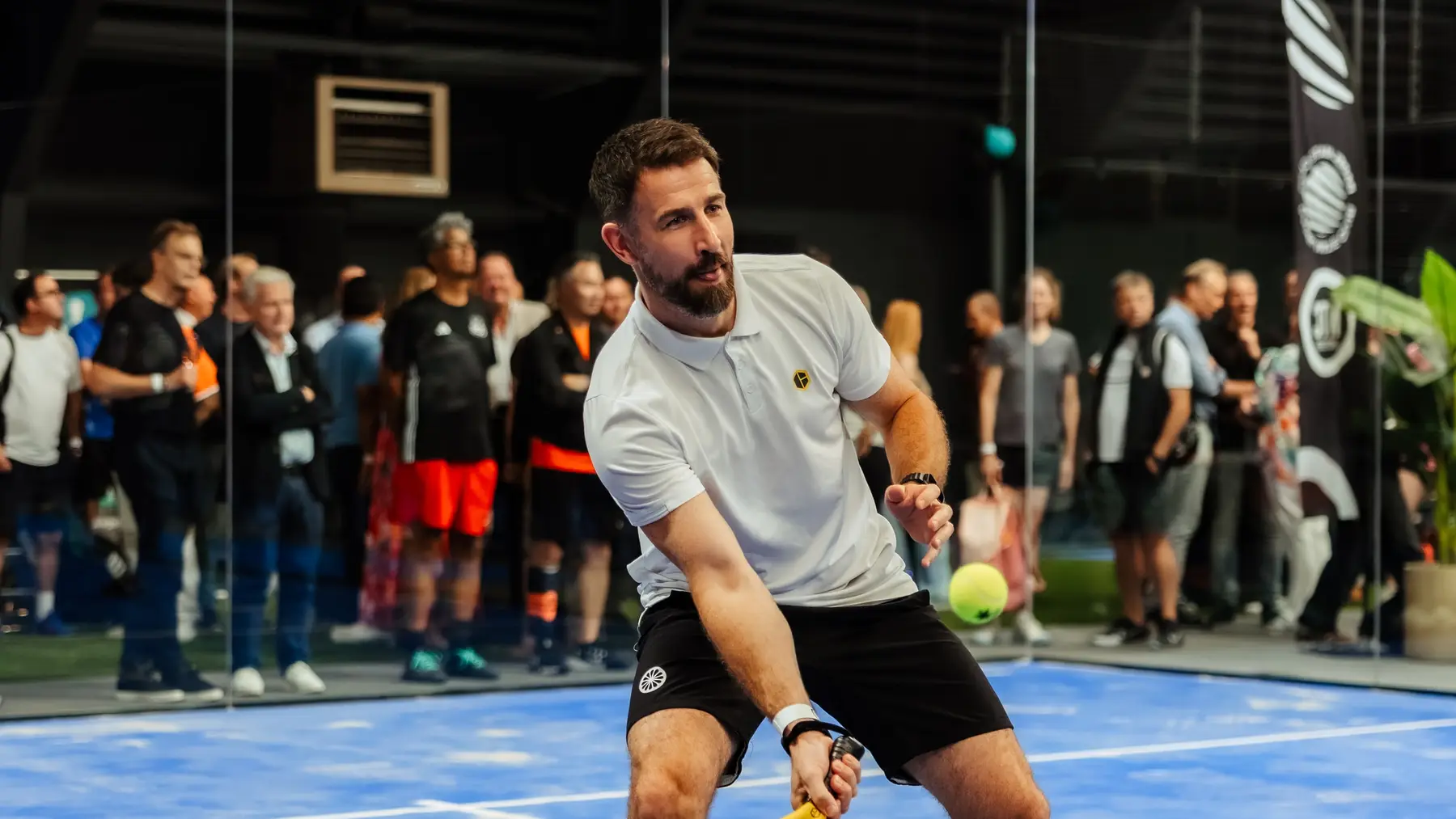 Een padelspeler speelt een bal voor een publiek dat vanaf de zijkant van de baan toekijkt.