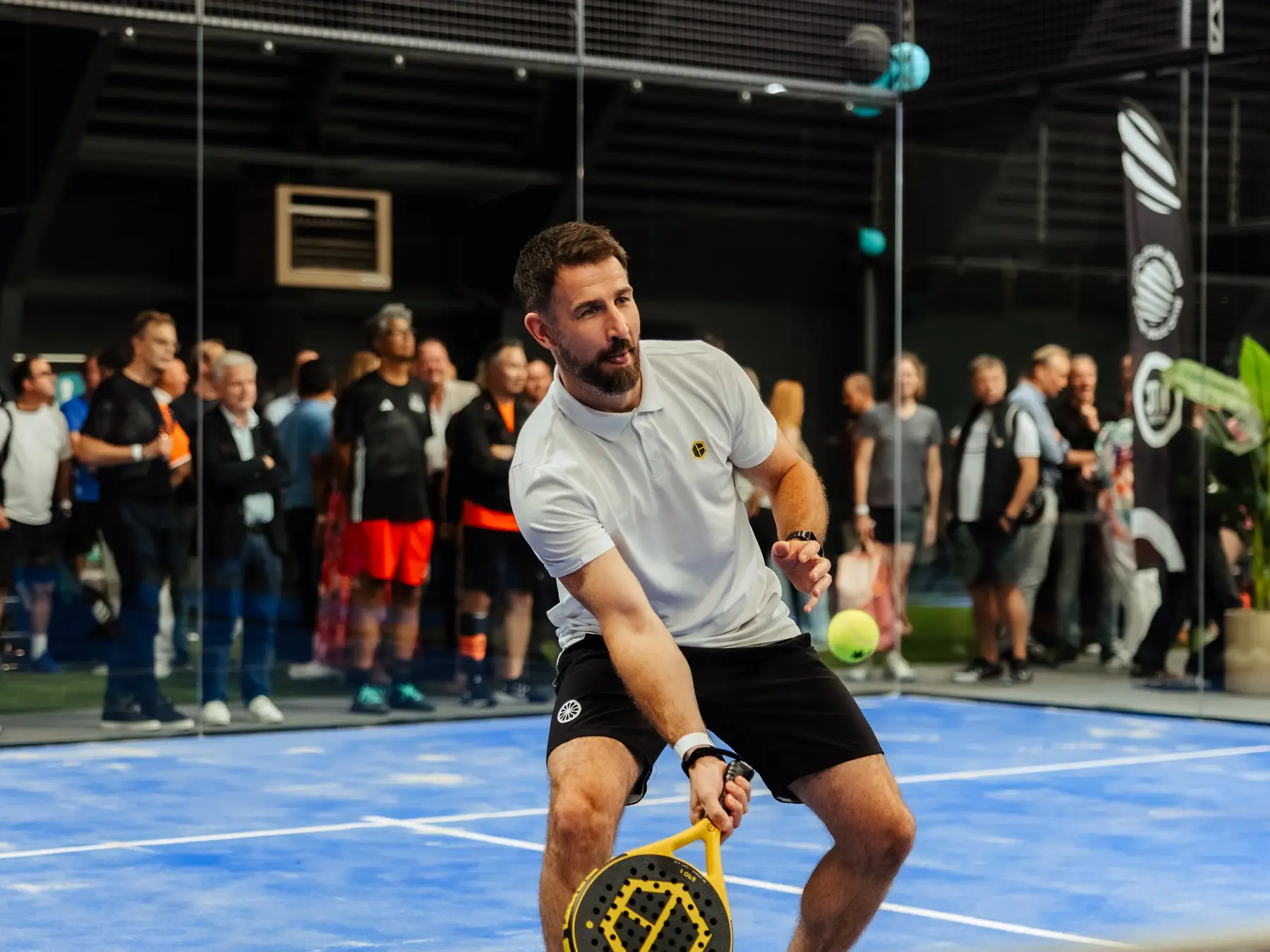 Een padelspeler speelt een bal voor een publiek dat vanaf de zijkant van de baan toekijkt.