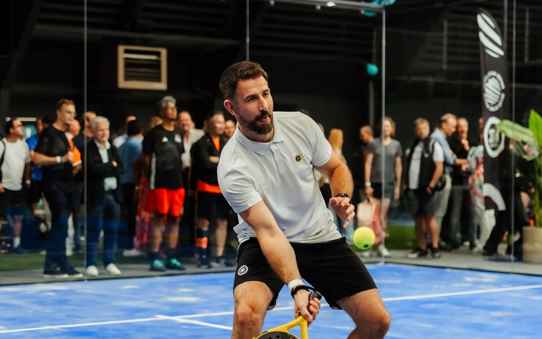 Een padelspeler speelt een bal voor een publiek dat vanaf de zijkant van de baan toekijkt.