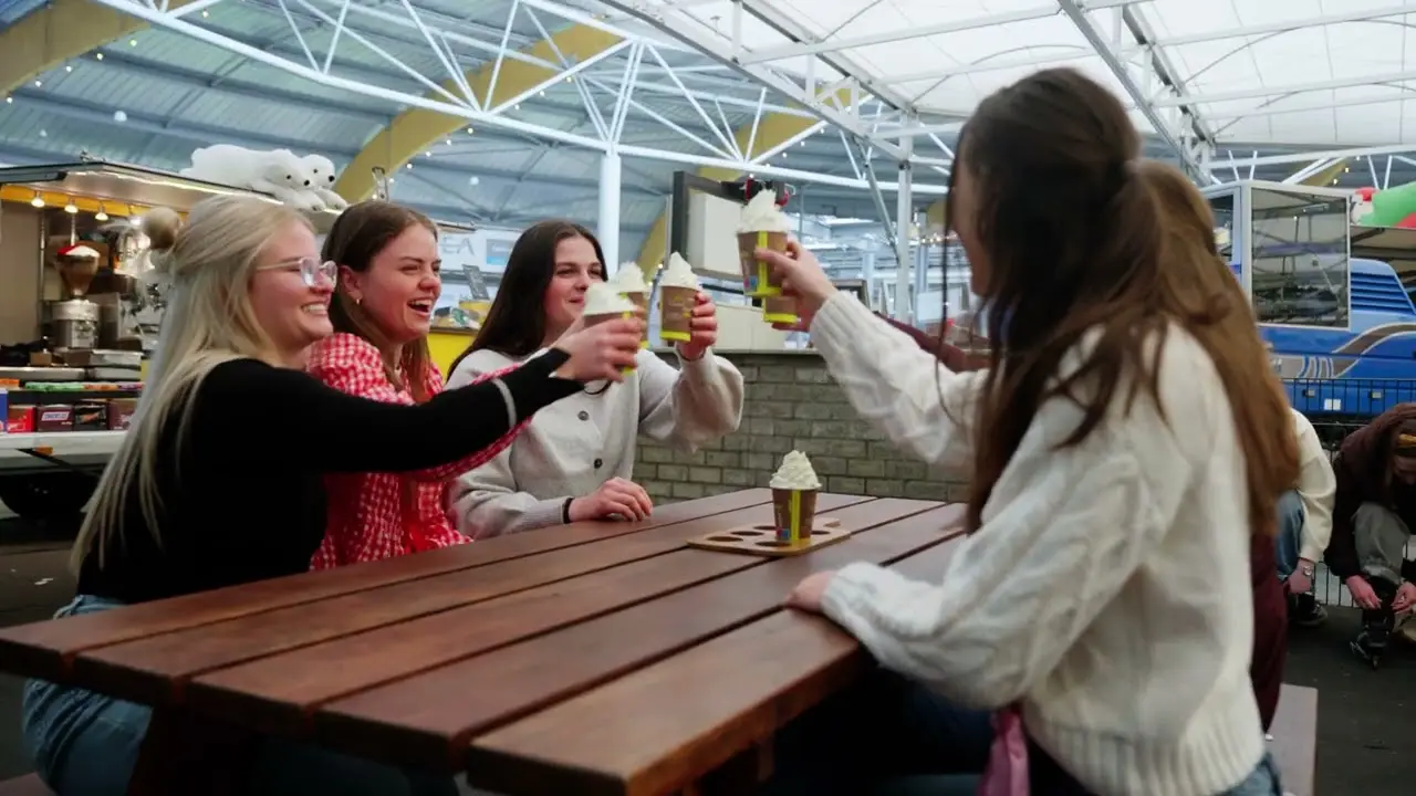 Vier jonge vrouwen proosten met warme chocolademelk aan een tafel bij de ijsbaan.