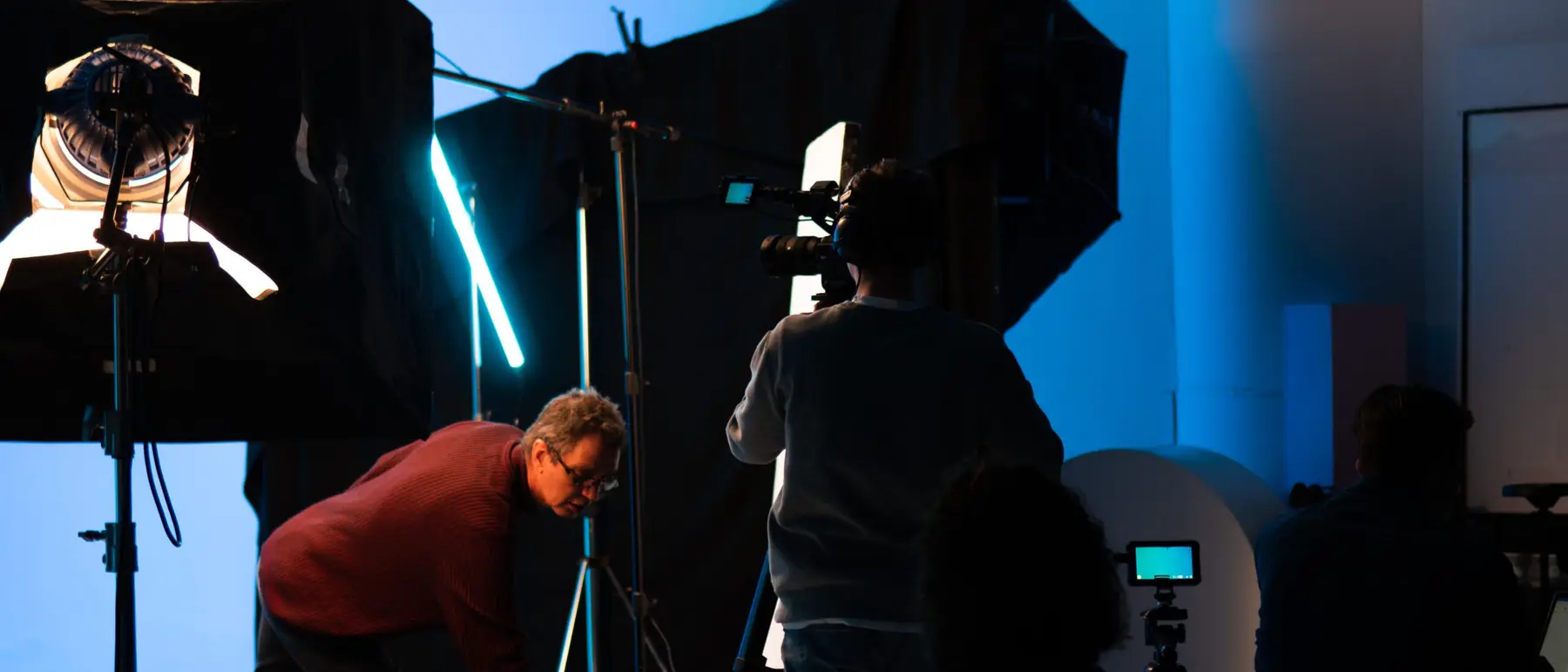 Achter de schermen bij een opname: lichtschermen, statieven en een crew tegen een blauwe achtergrond.