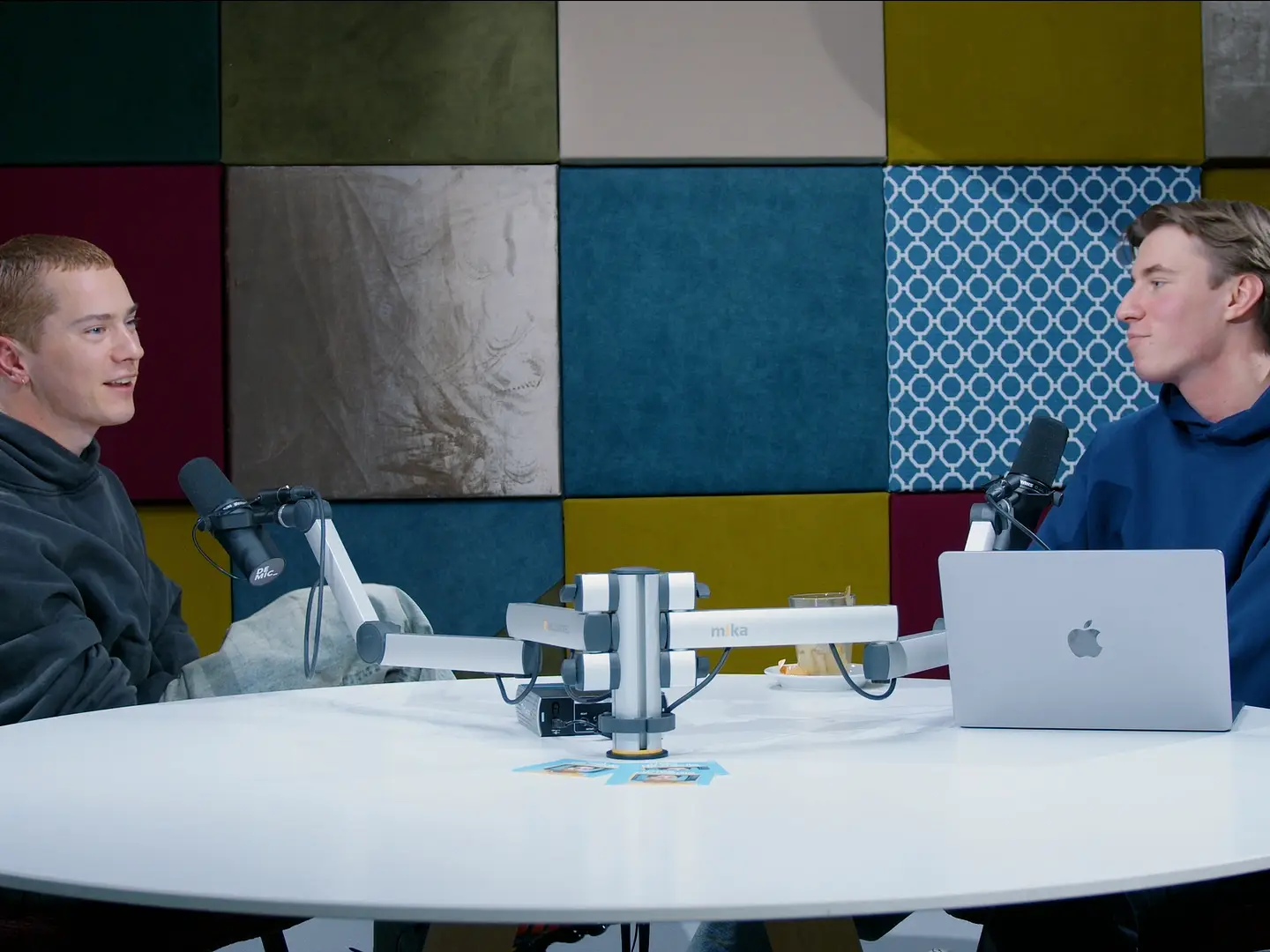 Twee mannen in hoodies in gesprek achter studiomicrofoons met een laptop op tafel.