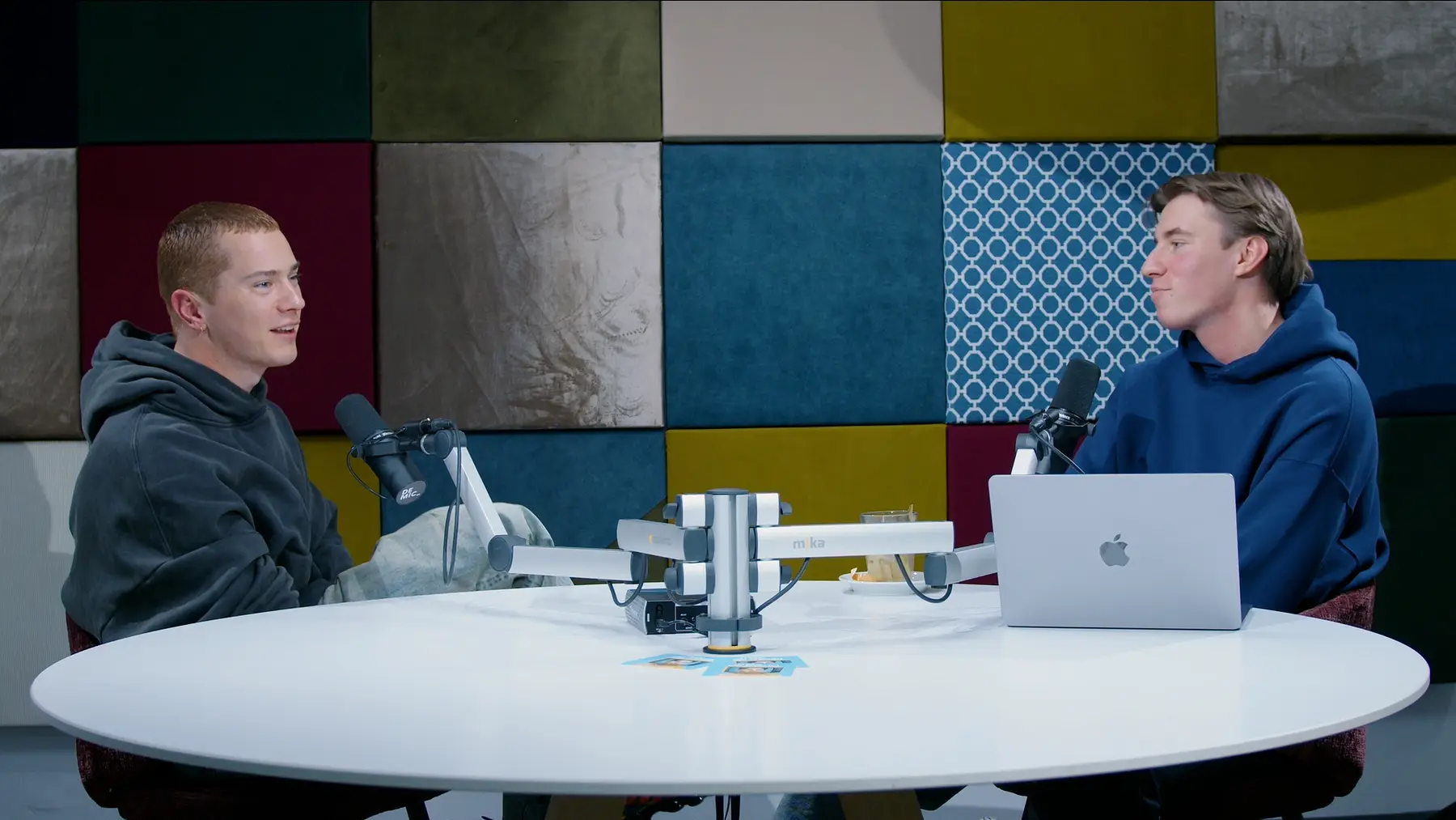 Twee mannen in hoodies in gesprek achter microfoons, met een laptop op tafel.