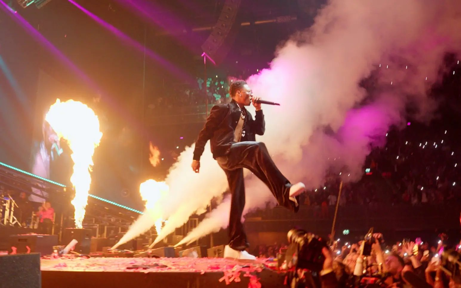 Een artiest springt over het podium tussen vuurzuilen en rook tijdens een concert.