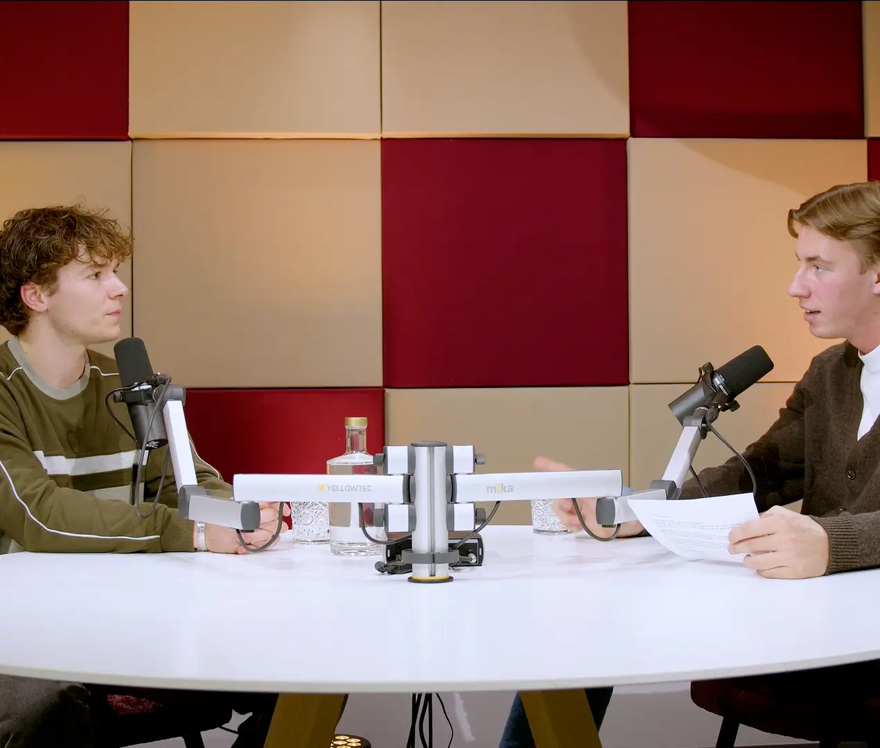 Twee jonge mannen in gesprek aan een ronde witte tafel met microfoons, voor kleurrijke akoestische panelen.