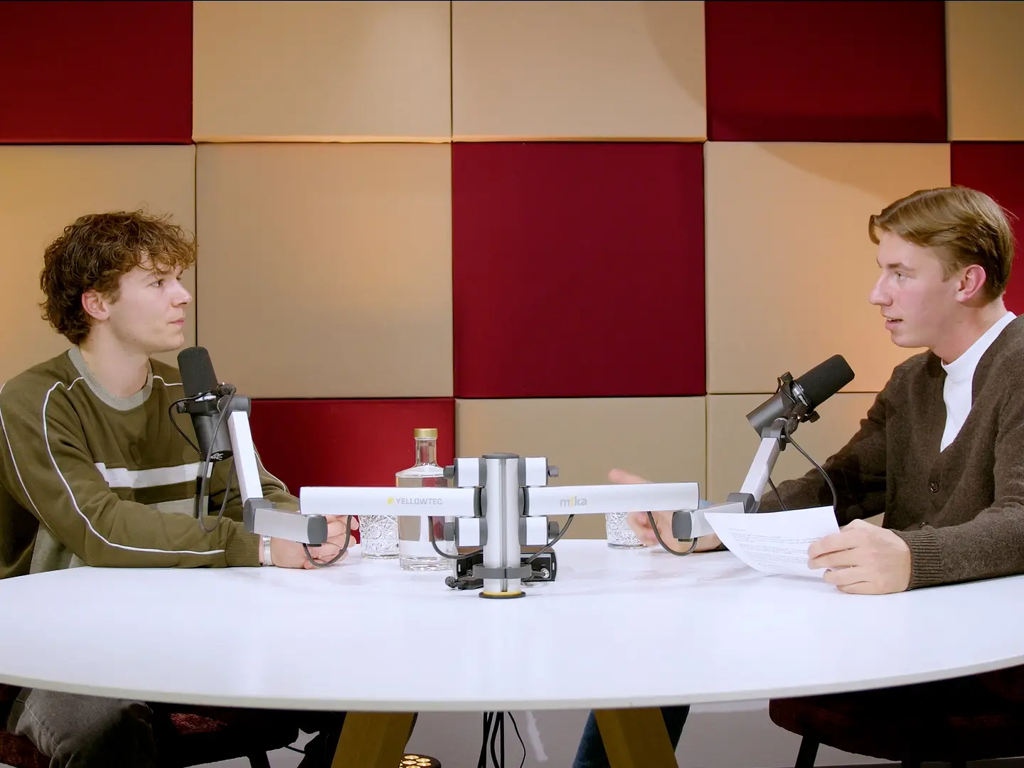 Twee jonge mannen in gesprek aan een ronde witte tafel met microfoons, voor kleurrijke akoestische panelen.