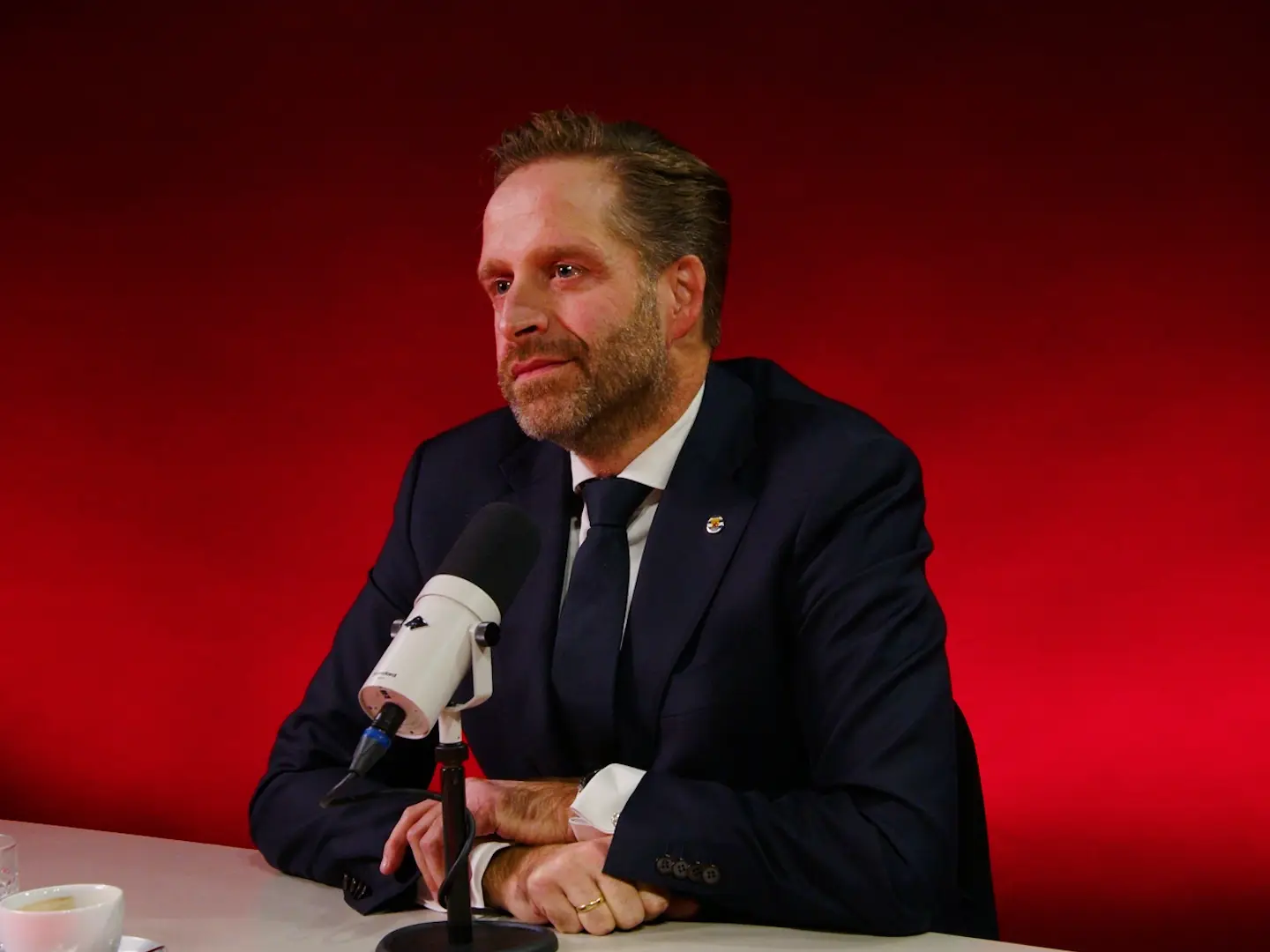 Een man in donker pak zit aan tafel achter een studiomicrofoon, tegen een rode achtergrond.