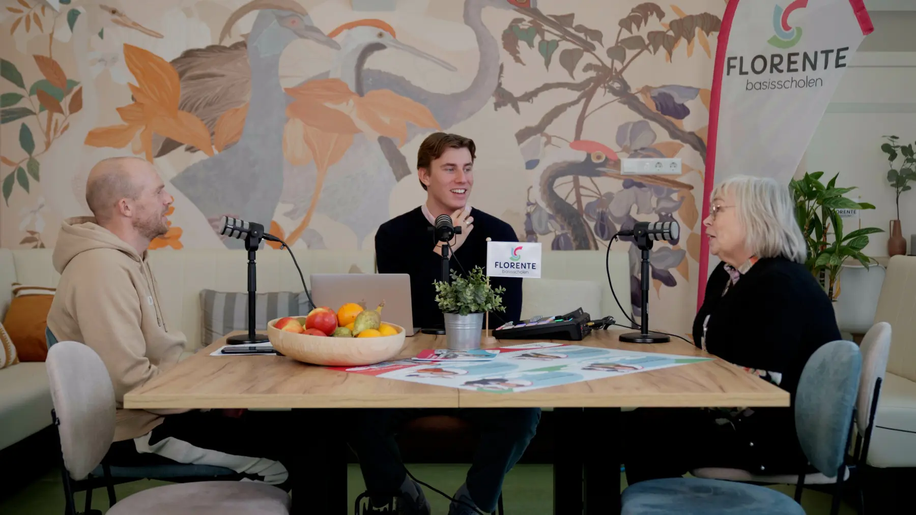 Drie mensen in gesprek aan een houten tafel met podcastmicrofoons, voor een kleurrijke muurschildering.