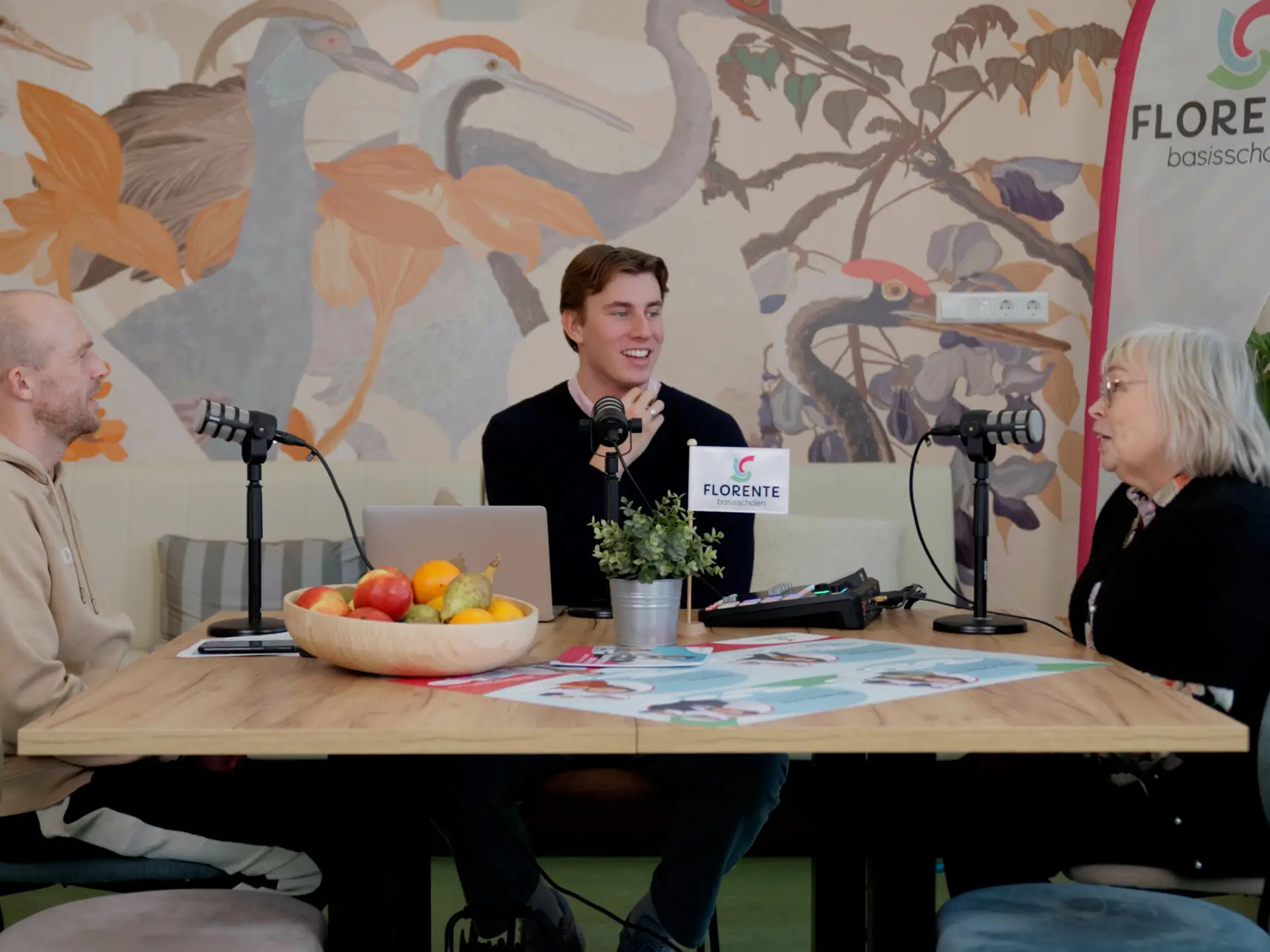 Drie mensen in gesprek aan een houten tafel met podcastmicrofoons en een fruitschaal, voor een kleurrijke muurschildering.
