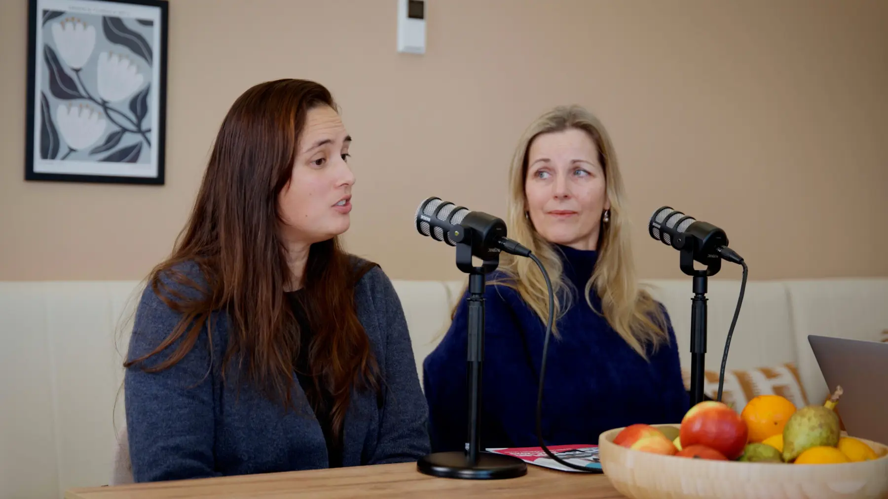 Twee vrouwen luisteren aan tafel achter podcastmicrofoons.