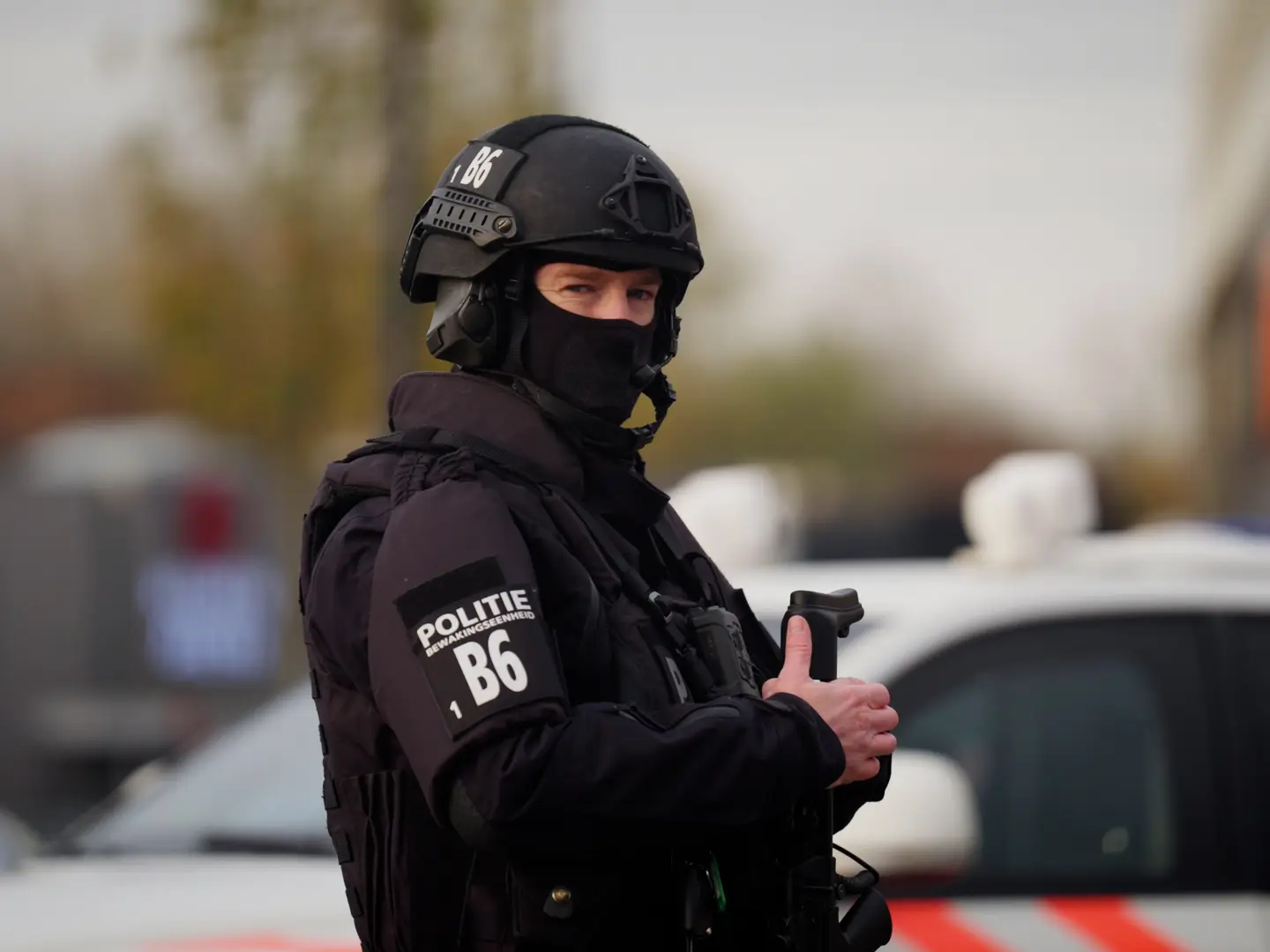Een zwaarbewapende agent van een arrestatieteam met bivakmuts staat bij een politieauto.