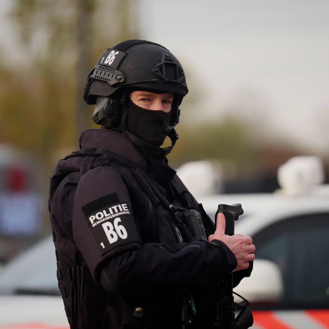 Een zwaarbewapende agent van een arrestatieteam met bivakmuts staat bij een politieauto.