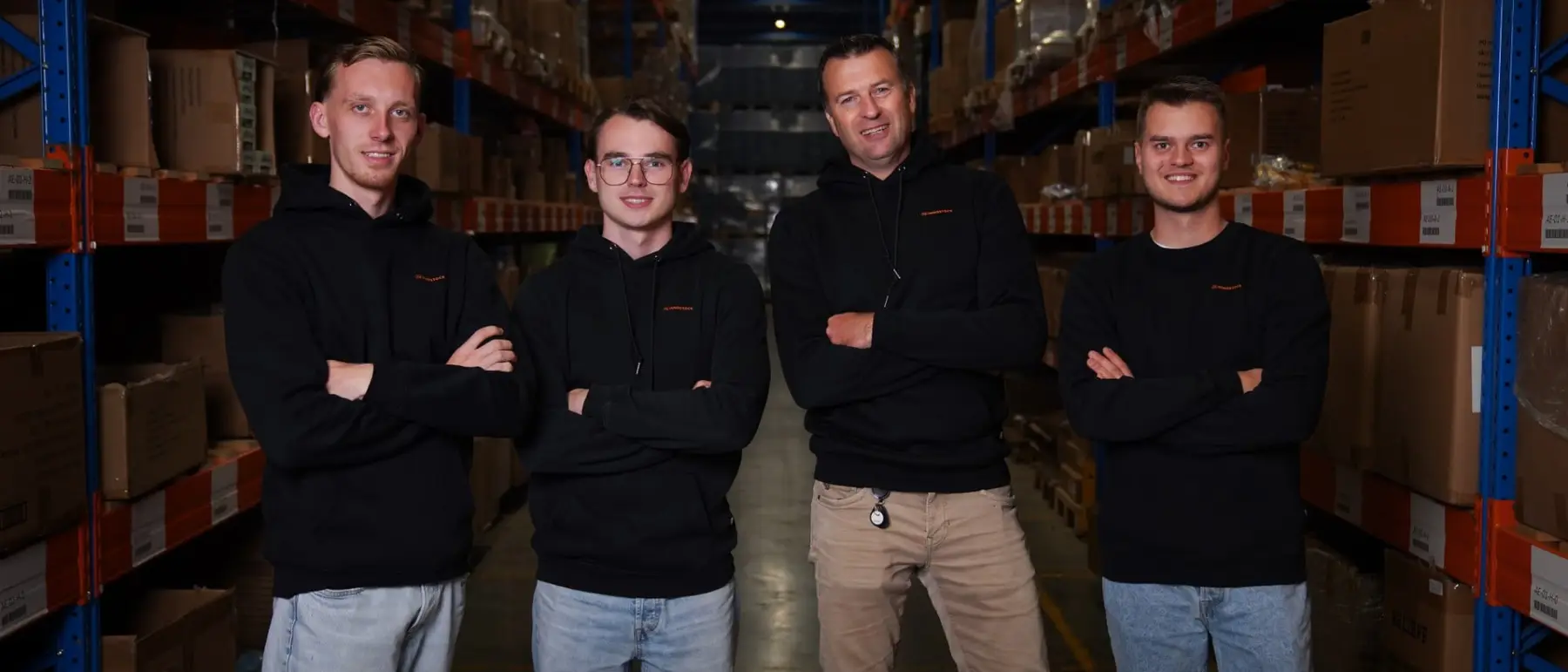 Vier medewerkers van Innostock in zwarte truien staan met de armen over elkaar in een gang tussen de stellingen van het magazijn.