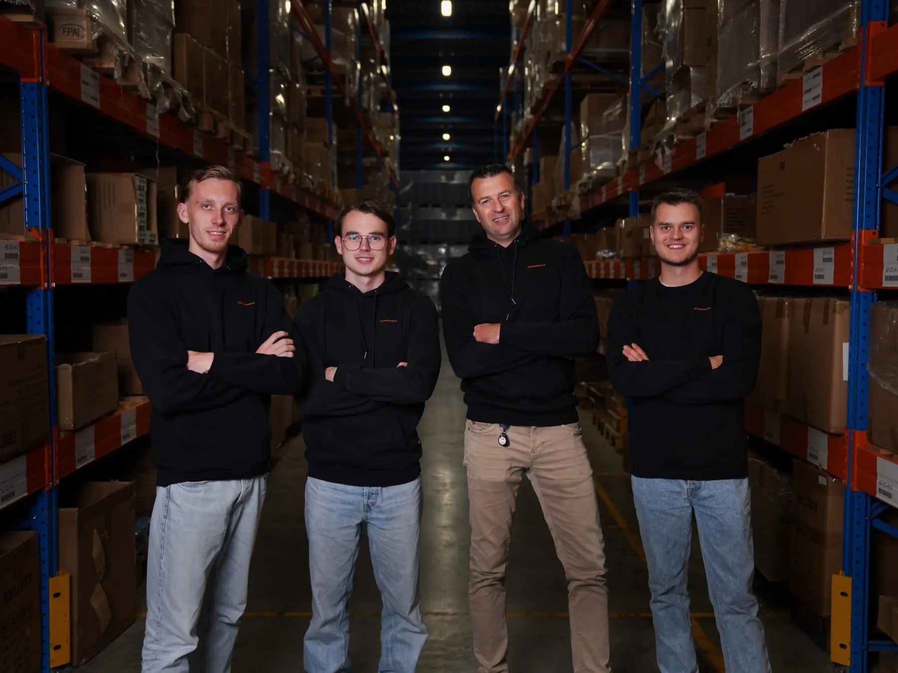 Vier medewerkers van Innostock in zwarte truien staan met de armen over elkaar in een gang tussen de stellingen van het magazijn.